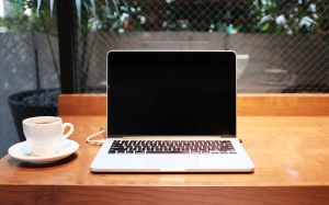 image of desk surface with coffee cup and saucer next to open laptop with blank screen - indicating people should check the website and social media to keep up with Twin Feathers' blog topics in 2024