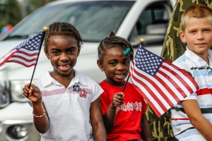 three children in front of white car, two holding an American flag