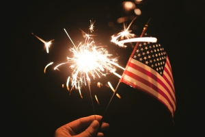 hand holding an American flag and sparkler against dark sky; 4th of July - why do we celebrate?